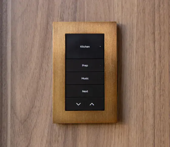 Control4 Lux keypad with black backlit buttons in a bronze-tone metal plate on a walnut wall